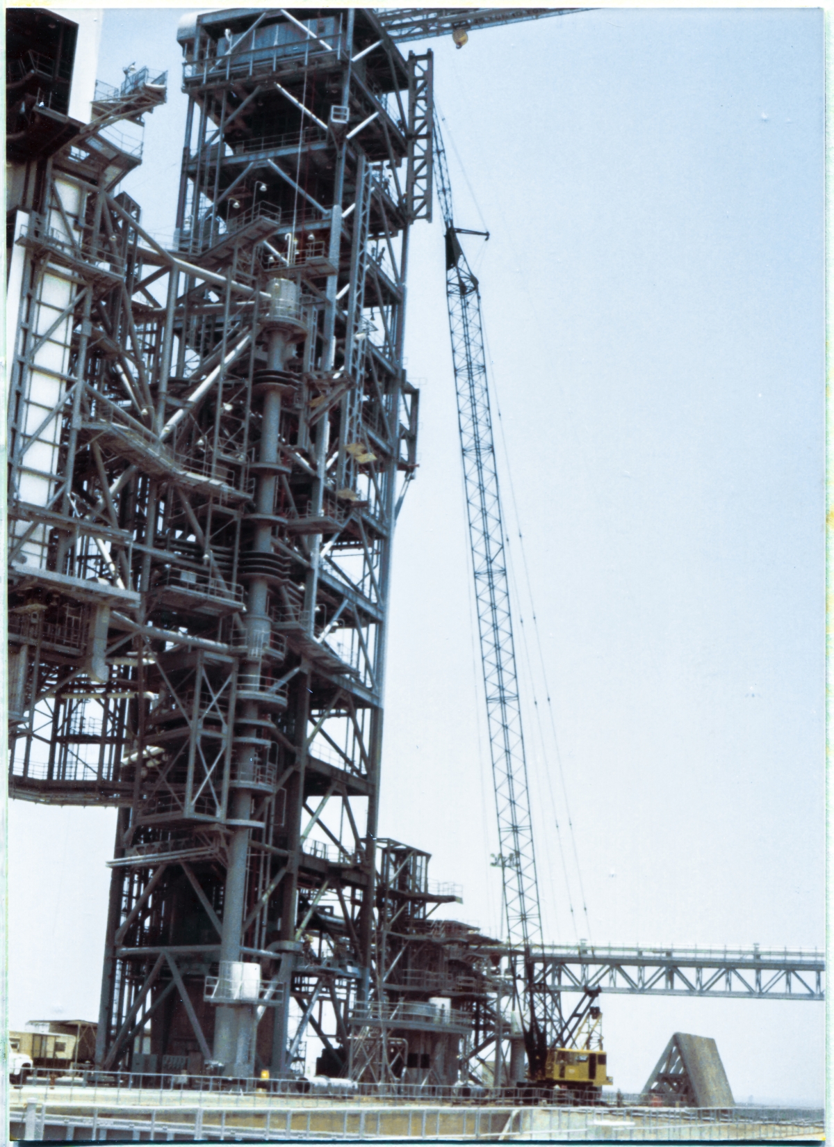 Image 080. The GOX Arm Strongback has reached its final elevation at the top of the Fixed Service Structure at Space Shuttle Launch Complex 39-B, Kennedy Space Center, Florida, with the tops of its twin W24x104 longitudinal members located just above elevation 300'-0”, which is the level of the deckplates on top of the FSS. The tag line which we saw earlier, attached to its lower right corner, was walked by the irownworker holding on to it, down on the ground, over to a position where it became draped across the front of the FSS, in direct contact with it, allowing other ironworkers up on the tower to reach it from up there, gather it in and get it out of the way, and continue using it to keep pulling the Strongback around, more or less to an orientation where you see it in this photograph. From there on, the tag line becomes unnecessary, is removed, and everything from here on is done with come-alongs, bringing much greater force to bear, getting the Strongback into its final position for welding and bolting it to the tower. As background, the colossal size of the FSS, spilling out of the frame up above, becomes apparent, and our once quite-large Strongback has now been dwarfed into insignificance by it. Photo by James MacLaren.